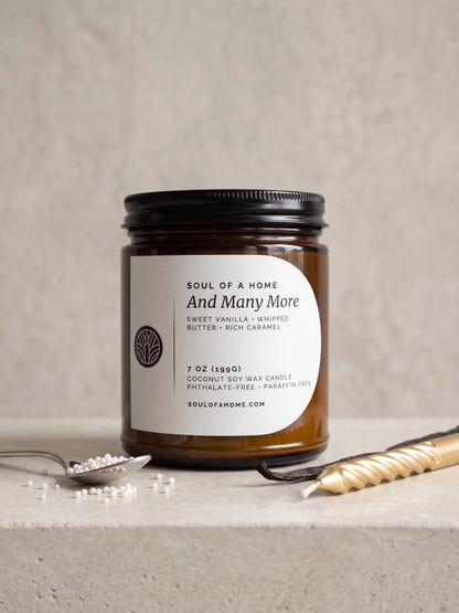A 7 oz soy wax candle in an amber glass jar with a black lid and a white label reading 'Soul of a Home And Many More', styled on a light stone surface with a spoon with cake sprinkles, a vanilla bean, and a gold birthday candle.