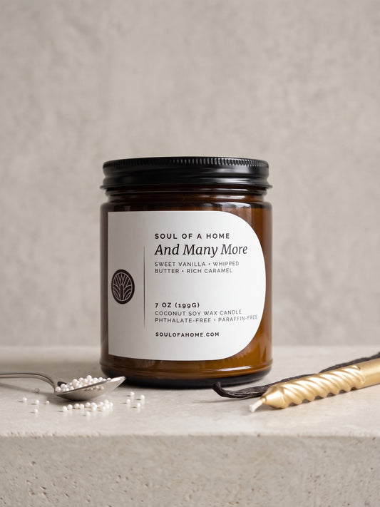 A 7 oz soy wax candle in an amber glass jar with a black lid and a white label reading 'Soul of a Home And Many More', styled on a light stone surface with a spoon with cake sprinkles, a vanilla bean, and a gold birthday candle.