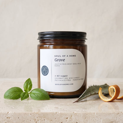 An amber glass jar candle with a black lid and a cream label reading 'Soul of a Home Grove' sits on a light stone surface with green basil leaves and a curled orange peel and aloe leaf around it .