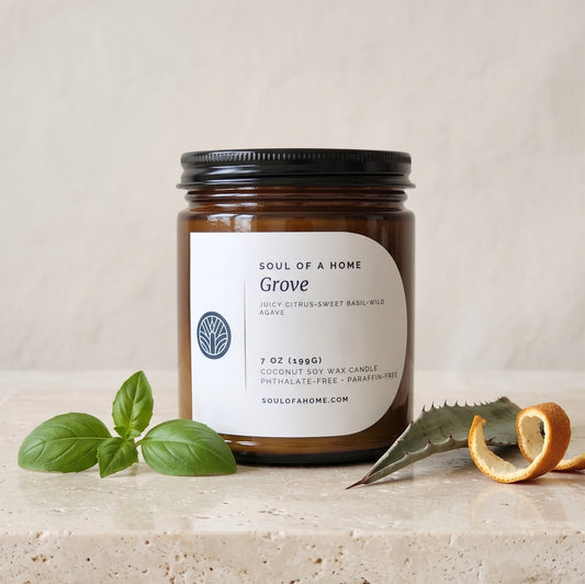 An amber glass jar candle with a black lid and a cream label reading 'Soul of a Home Grove' sits on a light stone surface with green basil leaves and a curled orange peel and aloe leaf around it .