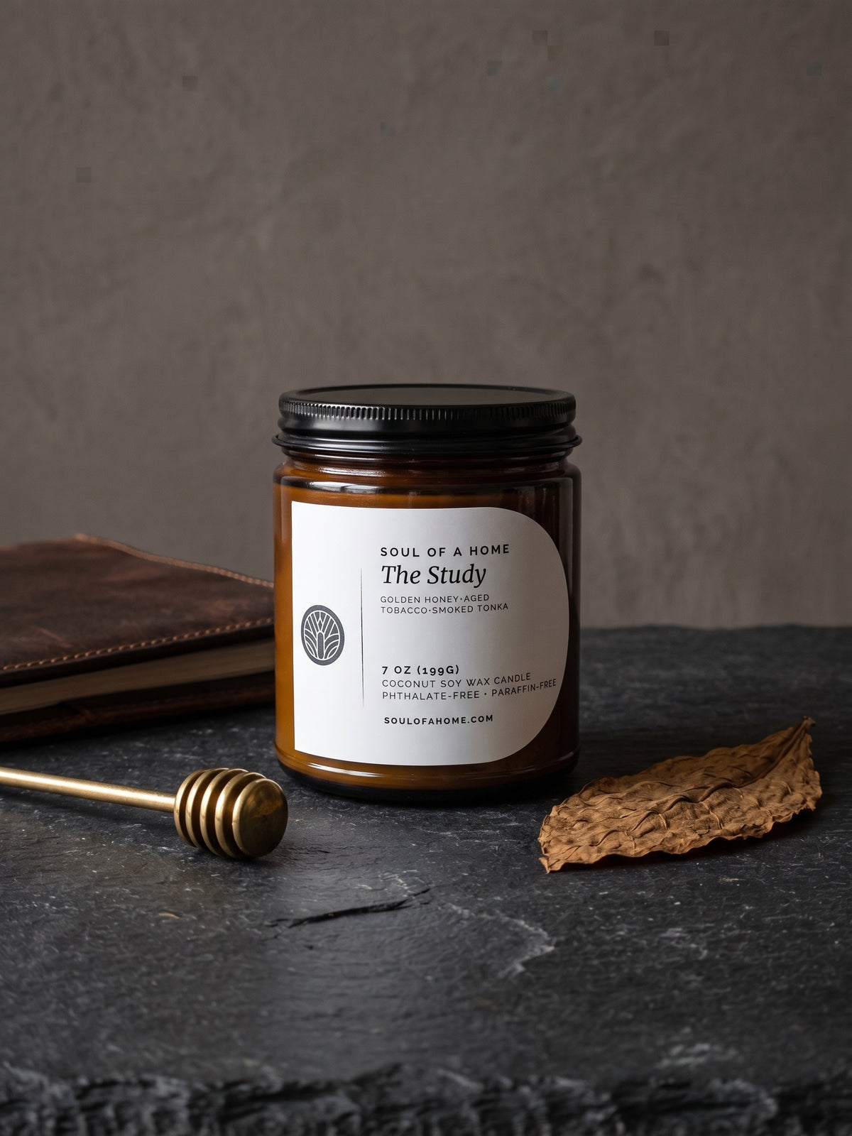 A 7 oz coconut soy wax candle in an amber glass jar with a black lid and a white label reading 'The Study' by Soul of a Home, styled on a dark slate surface with a honey dipper, a brown leather book, and a dried plant stem.