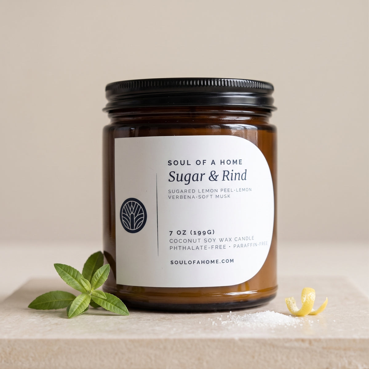 A 7 oz soy wax candle in an amber glass jar with a black metal lid and a white label reading 'Soul of a Home Sugar & Rind'. The candle is styled on a light stone surface with green verbena leaves and a small lemon rind against a warm beige background.