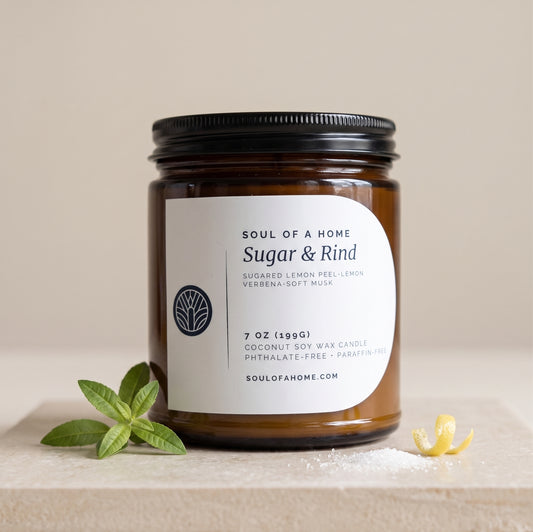 A 7 oz soy wax candle in an amber glass jar with a black metal lid and a white label reading 'Soul of a Home Sugar & Rind'. The candle is styled on a light stone surface with green verbena leaves and a small lemon rind against a warm beige background.