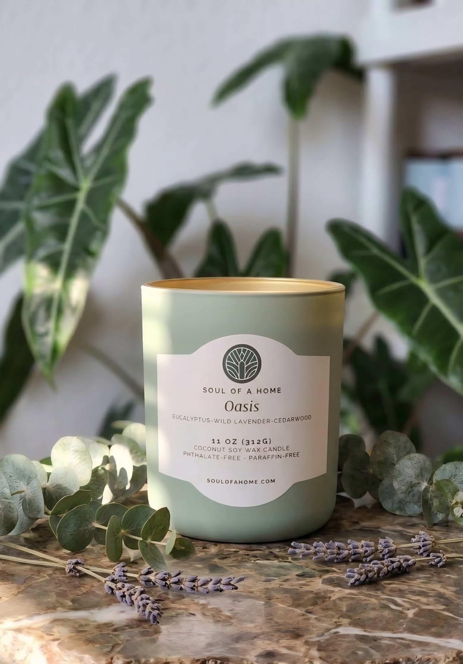 A sage green candle in a glass jar with no lid and a cream label reading 'Soul of a Home Oasis', styled on a marble surface with eucalyptus and lavender sprigs and large green leaves in the background.