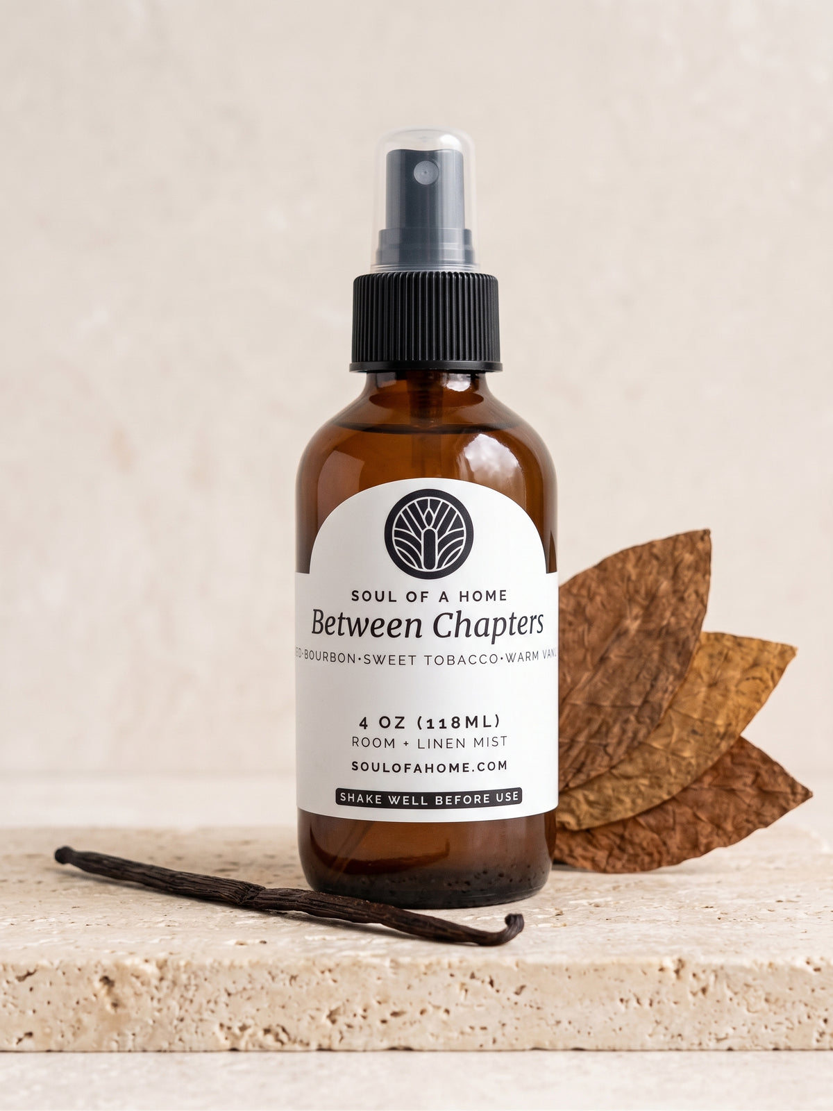 An amber glass room spray bottle with a black spray top and a white label reading 'Between Chapters' by Soul of a Home, styled on a stone surface with dried tobacco leaves and a vanilla bean.