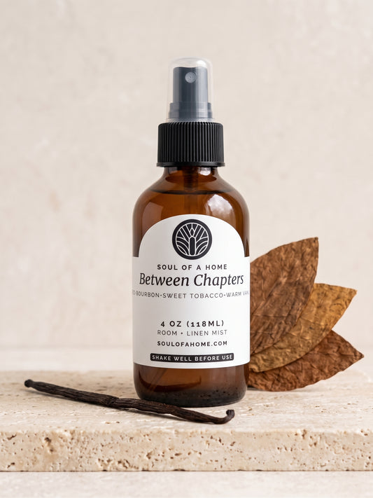 An amber glass room spray bottle with a black spray top and a white label reading 'Between Chapters' by Soul of a Home, styled on a stone surface with dried tobacco leaves and a vanilla bean.