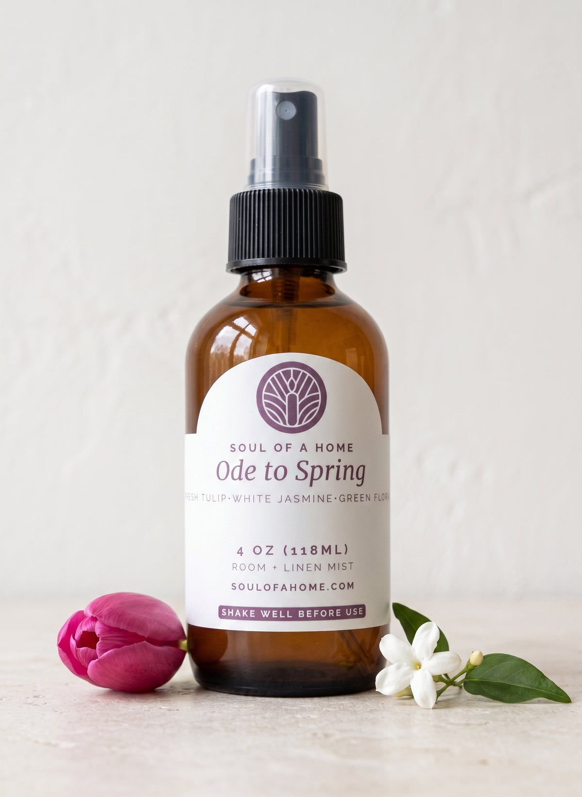 An amber glass room and linen mist spray bottle labeled 'Ode to Spring' by Soul of a Home, styled on a light surface with a pink tulip and a white jasmine flower.