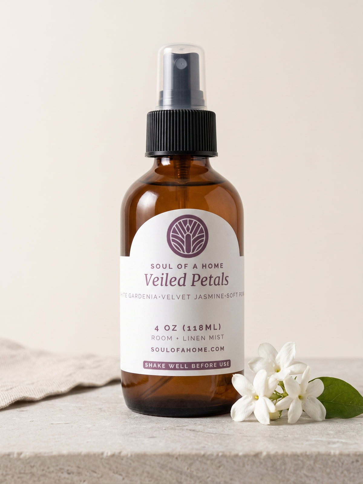 An amber glass room and linen mist spray bottle with a black spray top and a white label reading 'Soul of a Home Veiled Petals,' styled on a light stone surface with white jasmine flowers beside it.