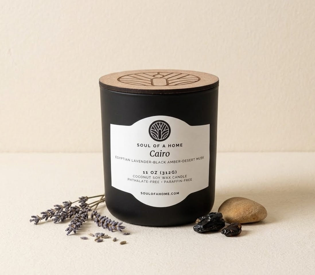 A black coconut soy wax candle jar with a light wood lid and a white label reading 'Soul of a Home' and 'Cairo,' styled on a cream surface with lavender sprigs, dark tonka, and a small stone.