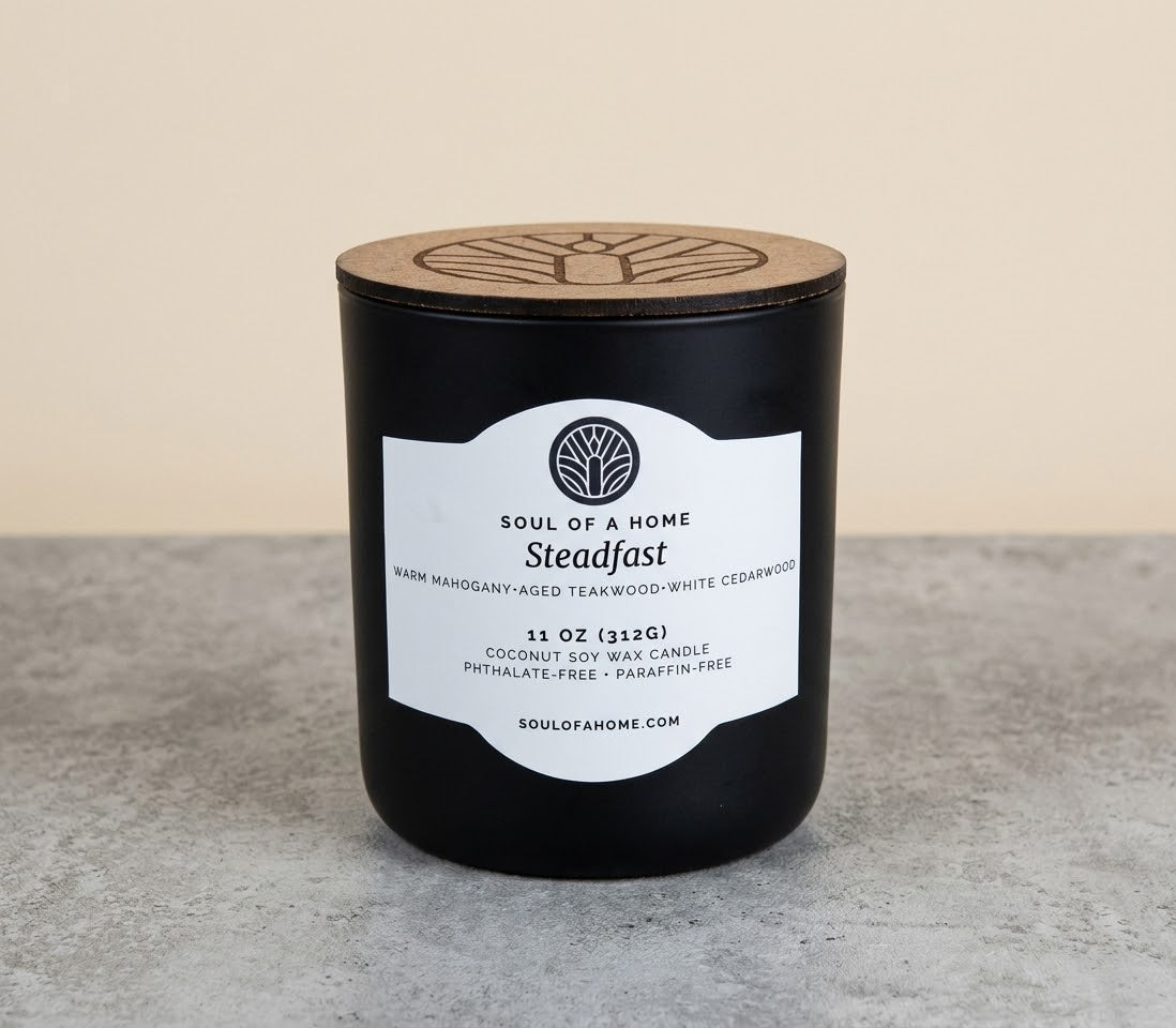 A black coconut soy wax candle jar with a light wood lid and a white label reading 'Soul of a Home Steadfast,' placed on a gray surface against a beige background.