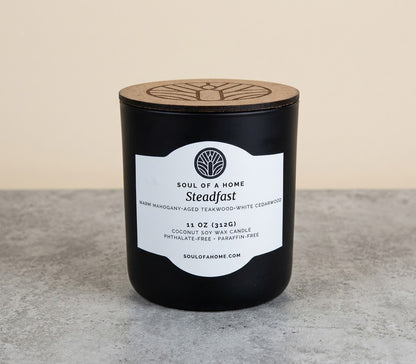 A black coconut soy wax candle jar with a light wood lid and a white label reading 'Soul of a Home Steadfast,' placed on a gray surface against a beige background.
