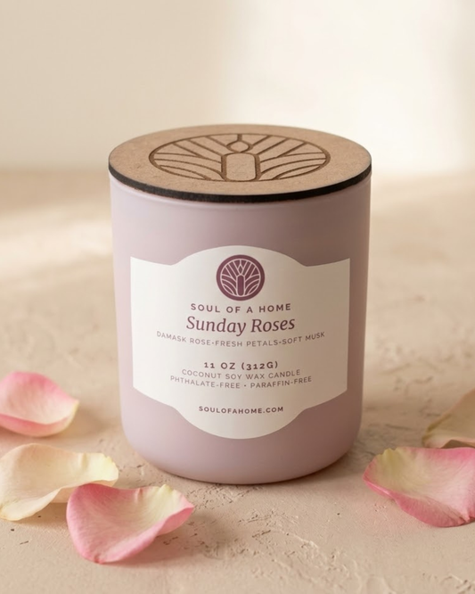 A coconut soy wax candle in a matte glass jar with a natural wood lid, labeled 'Sunday Roses' by Soul of a Home, styled with scattered pink rose petals on a beige surface.